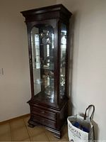 Full view of the tall solid mahogany display cabinet with glass door and sides placed against wall showing overall shape and finish.