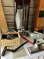 Kirby Heritage II upright vacuum cleaner with attached cloth bag, red wand/hose, additional parts and accessories inside a grid-patterned box placed beside it, and a gray accessory case