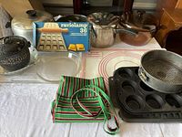Wide shot of pots, ravioli mold, muffin tins, pie dishes, and apron on table.