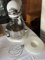 Front view of white KitchenAid Professional stand mixer with stainless steel mixing bowl and wire whip attachment and plastic cover on table
