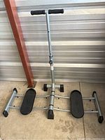 Full view of the foldable Leg Magic exercise machine, showing the silver metal frame, black foot pedals, and central vertical handlebar with black foam grips.