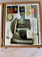 Microscope and accessories in foam insert inside wooden box.