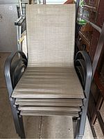 Four stacked outdoor patio chairs with beige sling seats and dark metal frames, showing armrest design and construction.