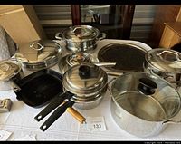 Wide shot showing seven pots and pans with lids on a table, highlighting variety of sizes and wooden handles on some cookware.