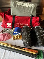 Full view of all sports equipment laid out on a bed including hockey bag, leg pads, gloves, pool cue, massage roller, and helmet
