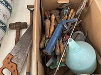 Box containing assorted hand and gardening tools showing wooden handles, metal blades, and a light blue funnel.