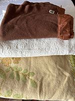 Photo showing folded brown wool bedspread with satin trim on top of a white textured bedspread and a green floral patterned bedspread beneath.