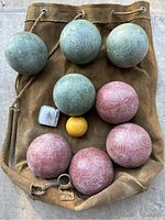 Top view of eight bocce balls (green and red/pink), small yellow pallino ball, metal measuring gauge, all on top of a brown canvas carrying bag with rope drawstring.