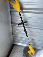Full view of the yellow Weed Eater electric string trimmer showing handle, shaft, and trimming head leaning against metal wall.