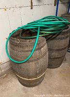Photo showing two wooden wine barrels with metal bands, placed side by side on a concrete floor, with a green garden hose coiled on top of one barrel.