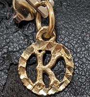 Close-up photo showing the 10K yellow gold letter 'K' pendant with textured circular frame and attached loop for chain.