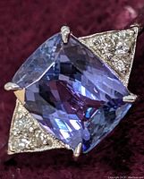 Close-up of cushion cut bluish-purple tanzanite set in four prongs between triangular diamond clusters on white gold band.