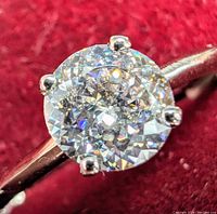 Close up image of round moissanite gemstone set in 10K white gold ring showing clear, sparkling facets and four prong setting on red velvet background.