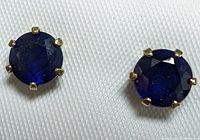 Front-facing close-up photo of the pair of round blue sapphire stud earrings set in 10K yellow gold prongs on a textured white surface.