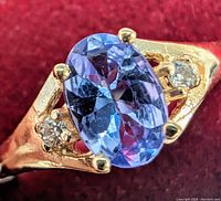 Close-up image of the ring showing the oval tanzanite center stone and the diamond accent stones on a red fabric background.