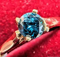 Close-up photo of the round blue diamond set in yellow gold ring, emphasizing the deep blue color and the prong setting.