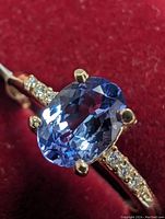 Close-up image showing the oval blue-violet tanzanite gemstone with yellow gold prong setting and pave diamond accents on the band.