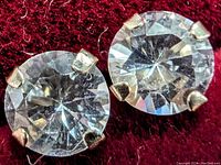 Close-up photo showing two round-cut white sapphire stones set in 14K yellow gold prong settings on a red background.