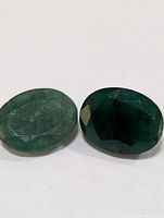 Two oval shaped emerald gemstones, one lighter green and one darker green, shown on a white background.