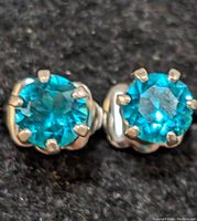 Close-up image of two silver stud earrings with round blue gemstones in prong settings on a black background.