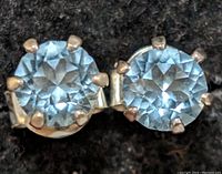 Close-up photo of one silver stud earring featuring a faceted blue round gemstone in a six-prong setting.