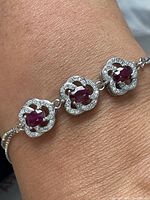 Close-up photo of the sterling silver bracelet worn on wrist showing three ruby-red oval stones each surrounded by clear cubic zirconia in floral-shaped silver settings.