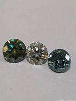 Three round cut moissanite stones showing color variations - blue-green and clear, placed on white background for clarity.