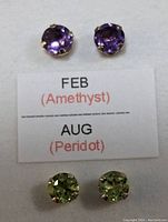 Two pairs of gemstone stud earrings pinned to a white card with printed card stating 'FEB (Amethyst)' and 'AUG (Peridot)'. Amethyst stones are purple, Peridot stones are light green.
