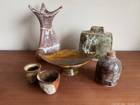 Photo showing all five ceramic pieces on a wooden surface against a plain wall: tall abstract vase, leaf-shaped bowl, square blossom-design vase, round corked bottle, and two small cups.