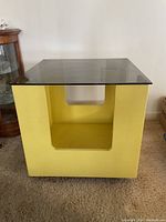 Front view of the yellow cube side table with clear glass top showing the hollow interior and cut-out design on the side.