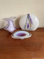 Three decorative glass pieces with purple accents including two vases and one bowl, photographed on wooden surface against wall.