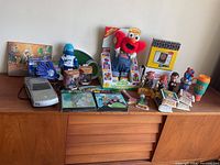 Wide view showing full lot including boxed Elmo plush, Pez dispensers, gray electronic device, sports foam finger, jars, mugs, and small collectibles.