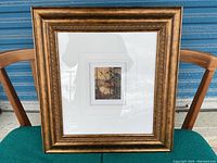 Full frontal view of framed art with landscape print inside ornate gold frame on a wooden chair against a blue background.