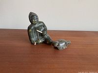 Photo showing two indigenous sculptures on a wooden surface, a kneeling person holding a needle and a smaller bird sculpture.