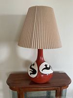 Front view of the red ceramic lamp with beige pleated shade, placed on a wooden side table.