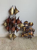 Two abstract metal wall art pieces leaning against a white wall on beige carpet. Each piece has layered geometric and organic metal shapes in warm metallic tones.