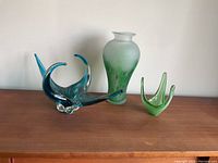 Set of three glassware pieces on wooden surface against white wall: blue decorative bowl, frosted green floral vase, small light green vase