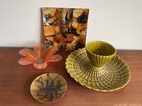 Photo showing orange glass flower dish, yellow ceramic basket and plate, small ceramic plate with abstract star design, and square decorative tile with abstract pattern arranged on wooden surface.