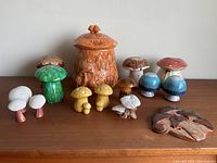 All items in the mushroom collection arranged on a wooden surface showing various ceramic mushroom figurines, the large lidded jar, and the mushroom wall hanging.