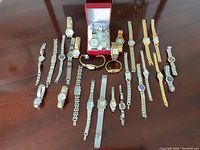 Photo showing 20 various wristwatches arranged on a wooden table, including metal link bands, mesh bands, and leather bands in silver and gold tones, with two watches in a red case.