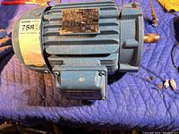 Blue electric motor placed on a padded blue surface showing the full side of the motor including the shaft and mounted electrical box. Has a label sticker with lot number on end.