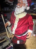 Santa Claus figure wearing red velvet suit with white faux fur trim, has white beard and glasses, mounted on red base on wooden surface with cluttered background.