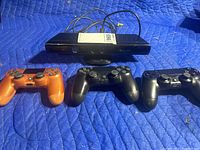 Photo showing three PlayStation DualShock 4 controllers in orange and black colors, and an Xbox Kinect sensor on blue quilted fabric.