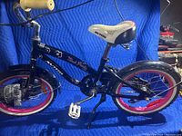 Side view of black girls youth bicycle AVIGO Black Pearl model with pink and white floral accents, red wheel rims, white padded seat, white pedals and training wheels on rear.