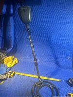 Full view of black metal detector with circular search coil on a blue stained padding surface alongside tools such as gloves and tape measure.