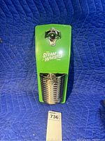 Front view of the green Steam Whistle branded bottle opener mounted on blue background, showing the attached silver cap collector basket with included mounting hardware.