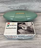 Closed green oval plastic case with gold Singer badge atop original buttonholer product box labeled 'Singer Buttonholer SIMS 4596'