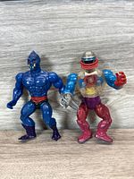 Front view of two Masters of the Universe action figures: a blue humanoid and a transparent robotic figure