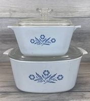 Two vintage Corningware baking dishes stacked. Blue cornflower design on white dishes. Clear glass lids with knob handles.