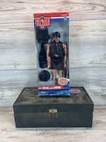 Photo of the GI Joe action figure in packaging standing on top of the wooden footlocker box on wood floor background.